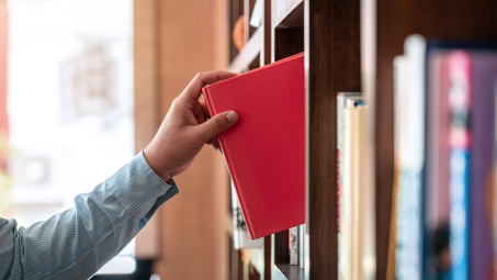 A close-up photograph of a person's hand pulling a red book from a crowded bookshelf.