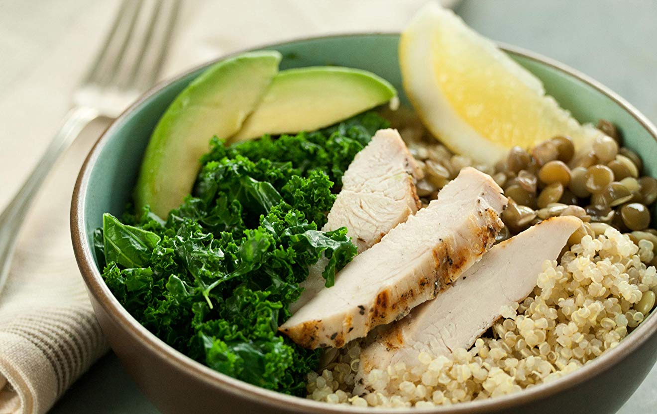 Image of protein bowl with quinoa, chicken, avocado, kale and lentils, garnished with lemon.