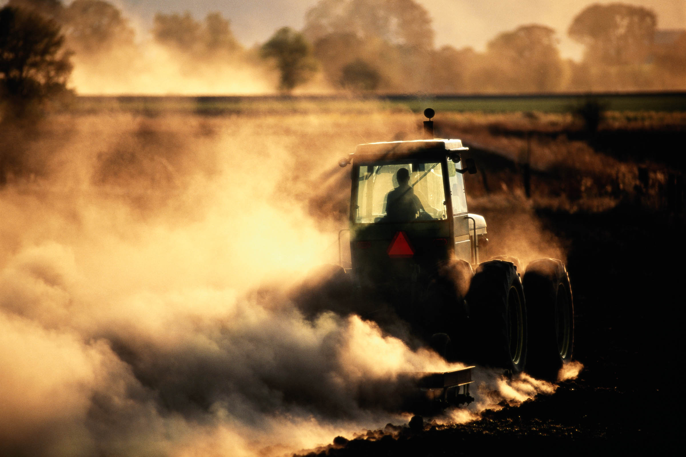 A hazy view of a tractor plowing a field surrounded by a dust cloud