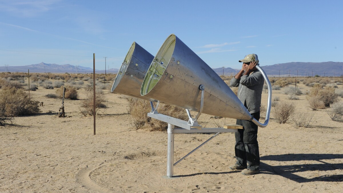 Two large metal cones on a stand in the desert, pointing outward, while a man holds a hollow pole that connects to the cones up to his ear