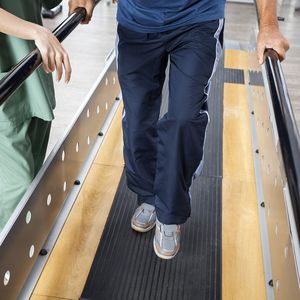 Senior Man Walking With Support Of Bars By Female Physiotherapis