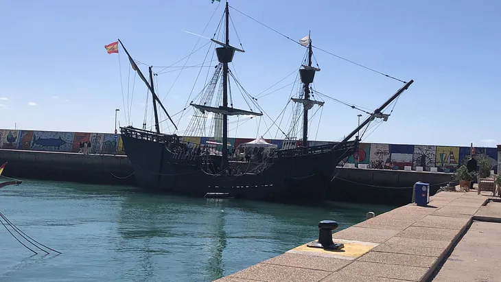 A Carrack ship docked in a harbour, flying the Spanish flag