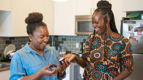 Two women in a kitchen laughing as they hold a jar of shea butter