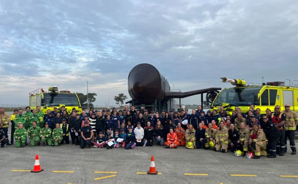 A group of about 100 volunteers and trainer posing in front of fire trucks and an aircraft replica used for firefighting training