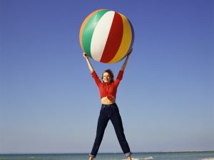 woman in pants and red shirt jumping up holding a beach ball on beach