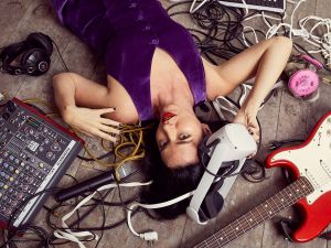 A woman with dark brown hair lays on the floor surrounded by an electric guitar and other musical instruments.