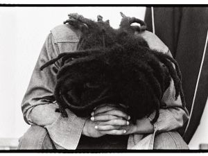 A man with dreadlocks leans down with hands clasped in a black and white photo
