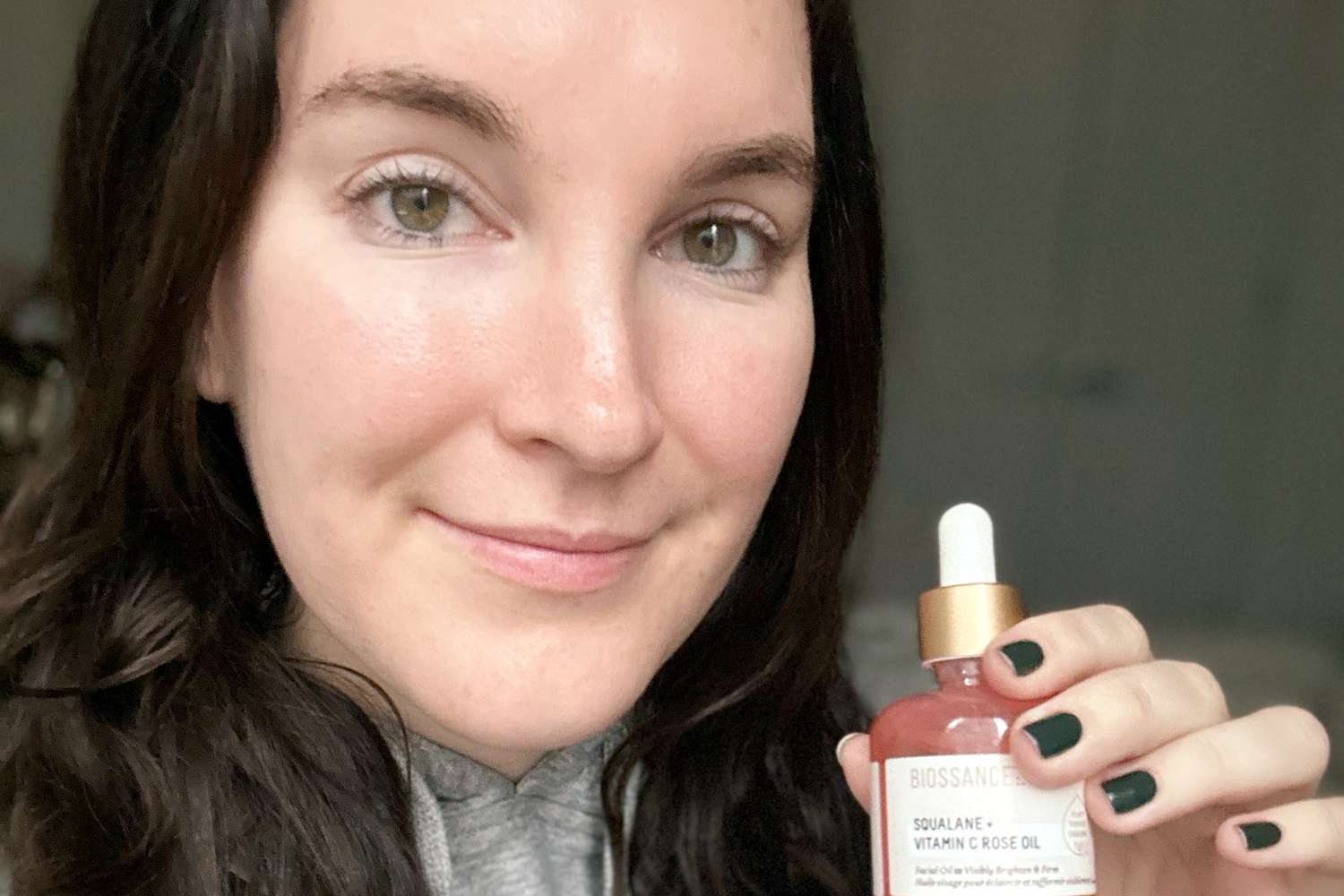 Person smiling and holding Biossance Squalane + Vitamin C Rose Firming Oil next to face