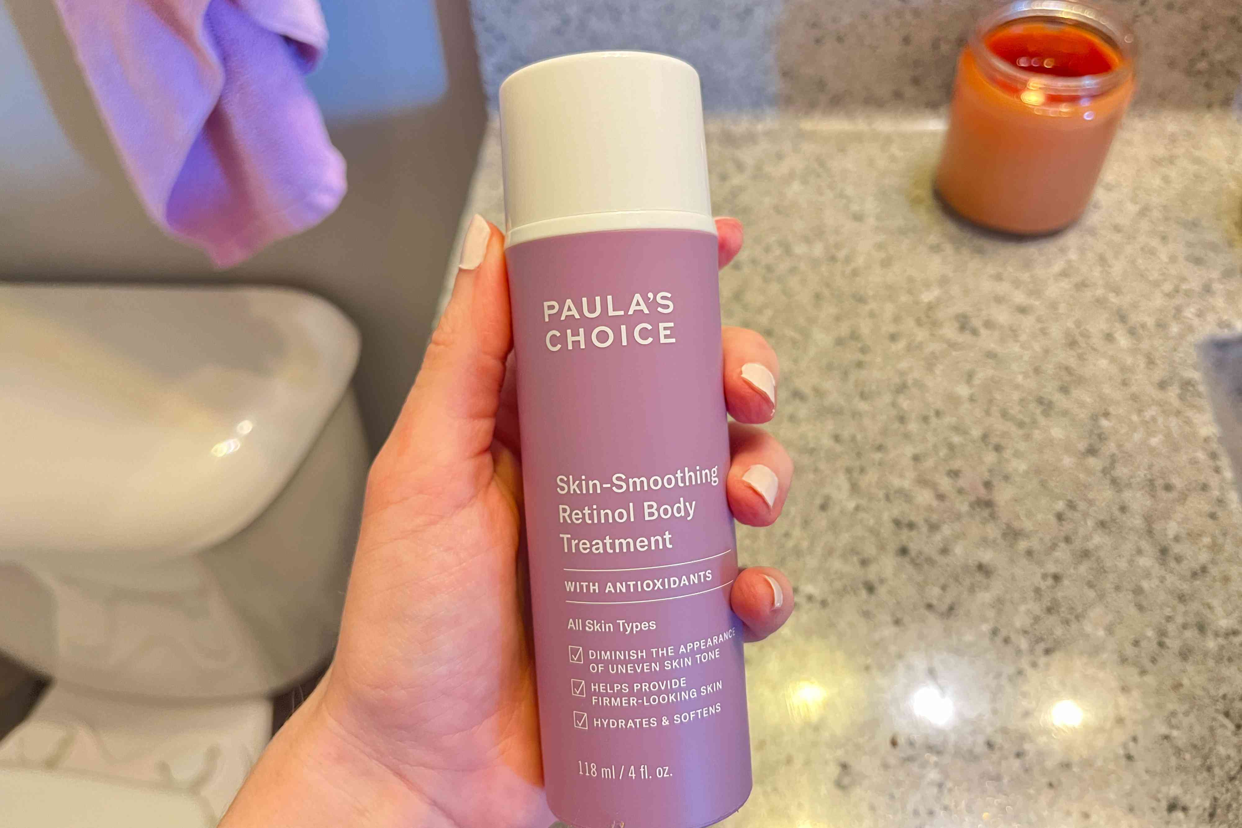 A person holds up a bottle of Paula's Choice Retinol Skin-Smoothing Body Treatment Shea Butter, Vitamin C & E Lotion, Moisturizer for Discoloration