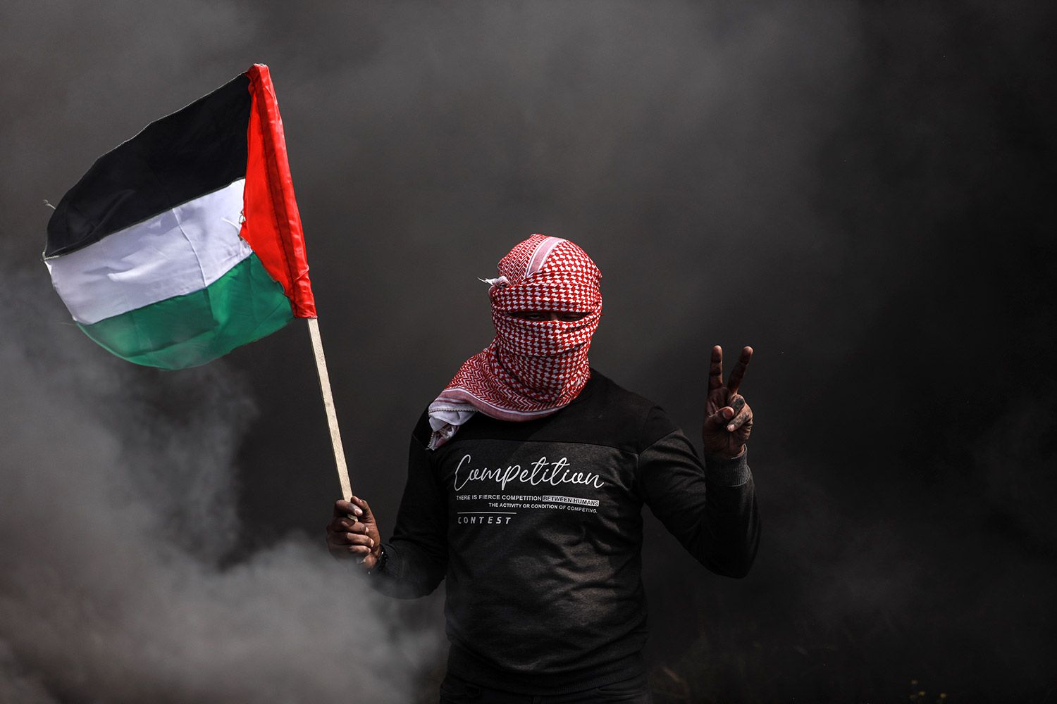 Figure with a covered face waves a Palestinian flag in front of a cloud of dark smoke