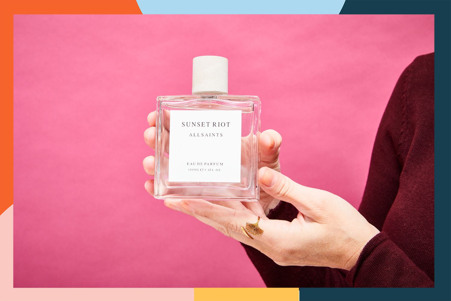 Hands holding the All Saints Sunset Riot Eau de Parfum in front of a pink background.