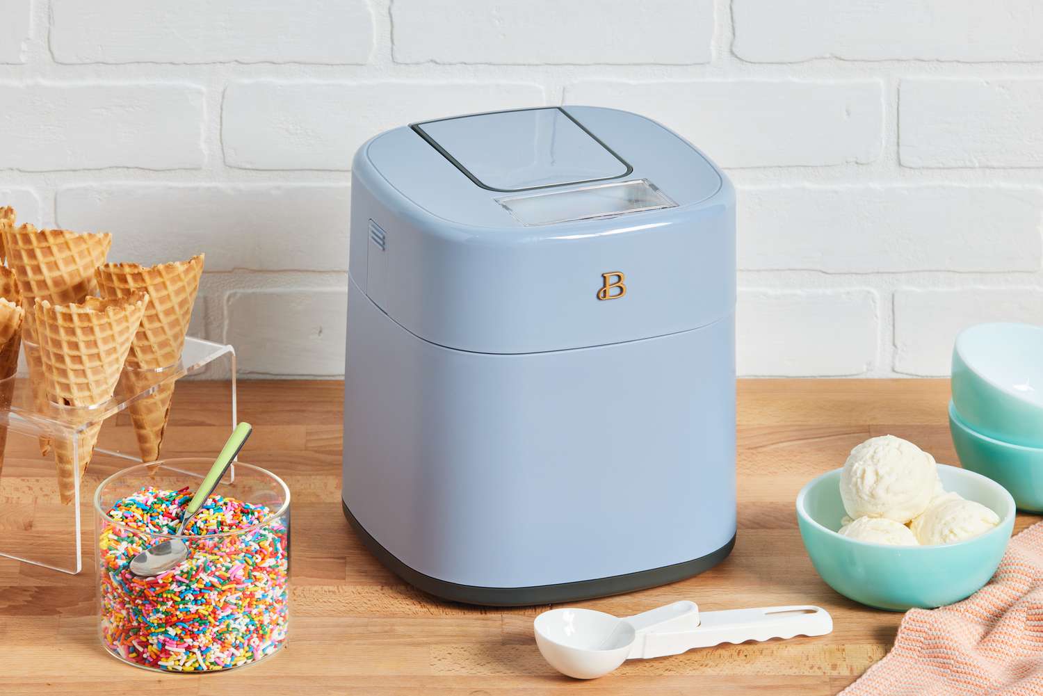 Beautiful by Drew Barrymore Ice Cream Maker sitting on a counter next to ice cream, sprinkles, and cones