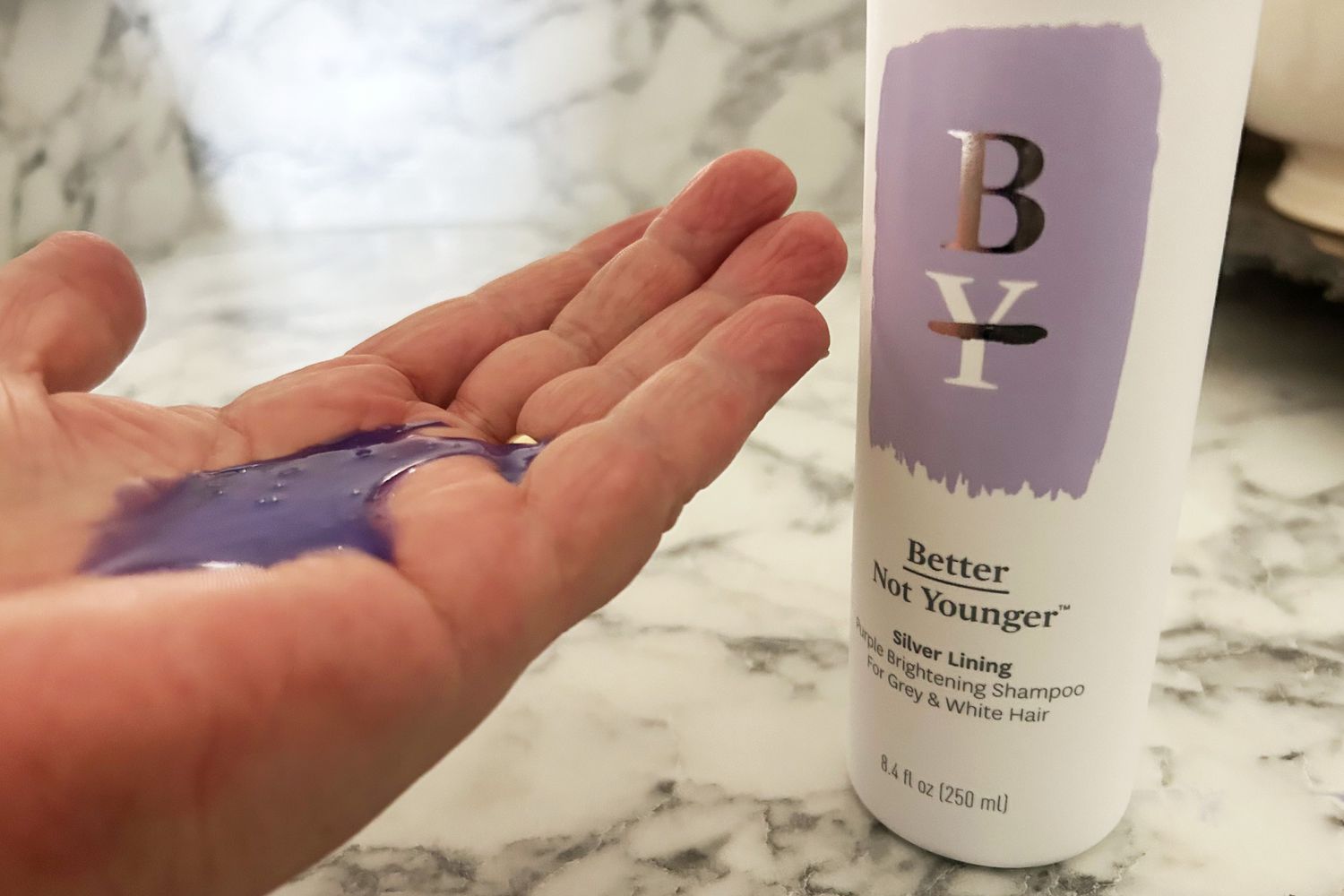 Hand with purple Better Not Younger Silver Lining Purple Brightening Shampoo for Grey & White Hair next to the bottle 