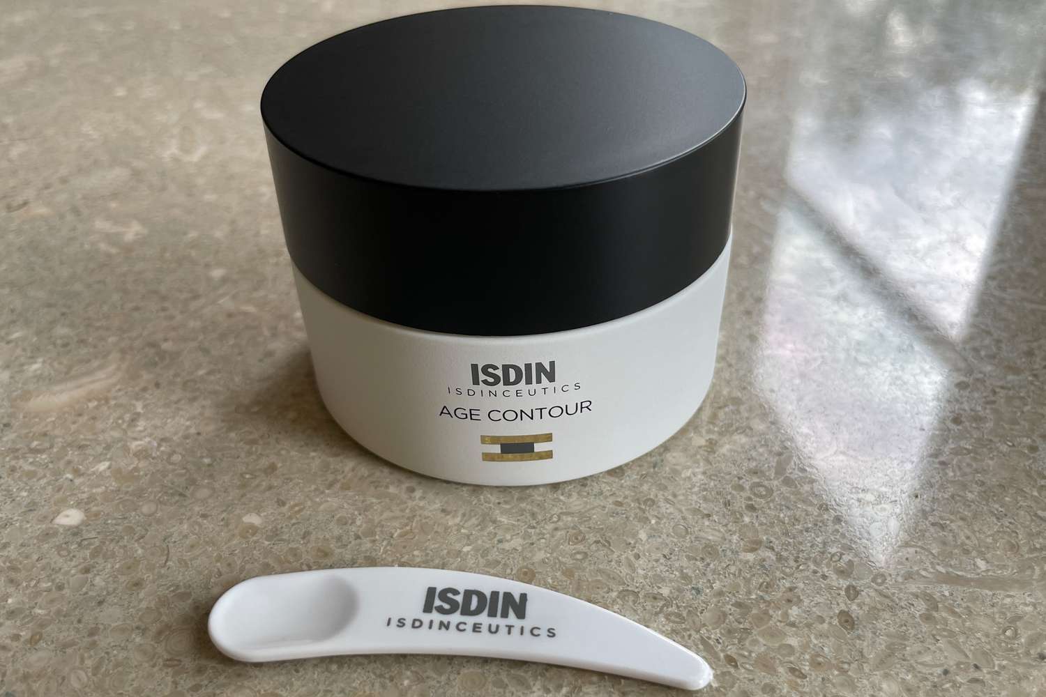 A jar of ISDIN ISDINCEUTICS Age Contour Firming and Rejuvenating Cream behind an applicator spoon