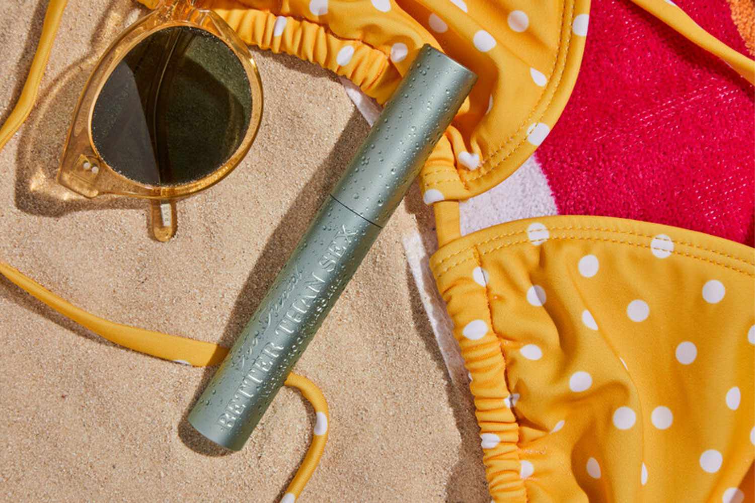 Too Faced Better Than Sex Waterproof Mascara next to a bikini top and sunglasses on sand