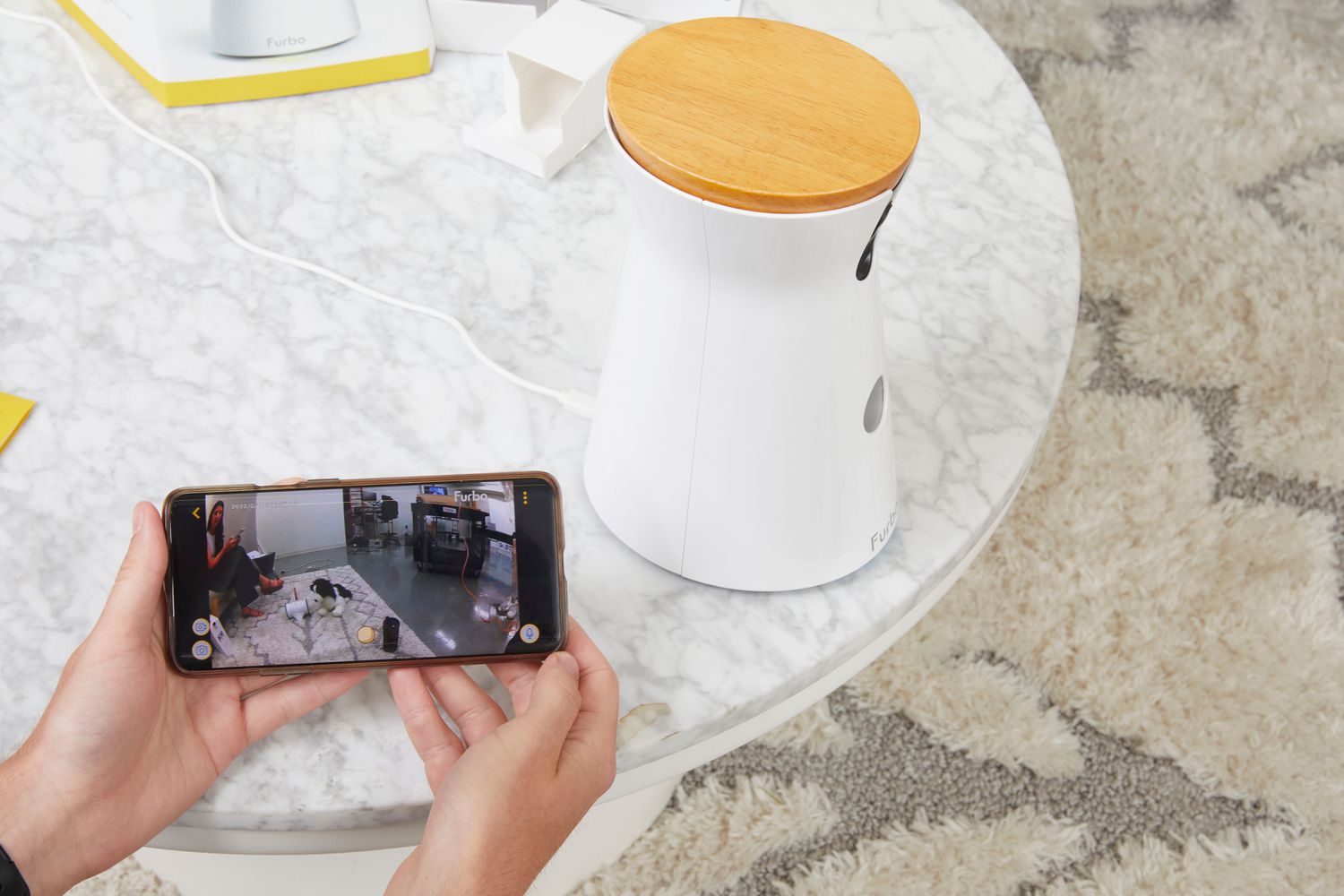 Hands holding smartphone while viewing video on app from Furbo 360° Dog Camera displayed on table