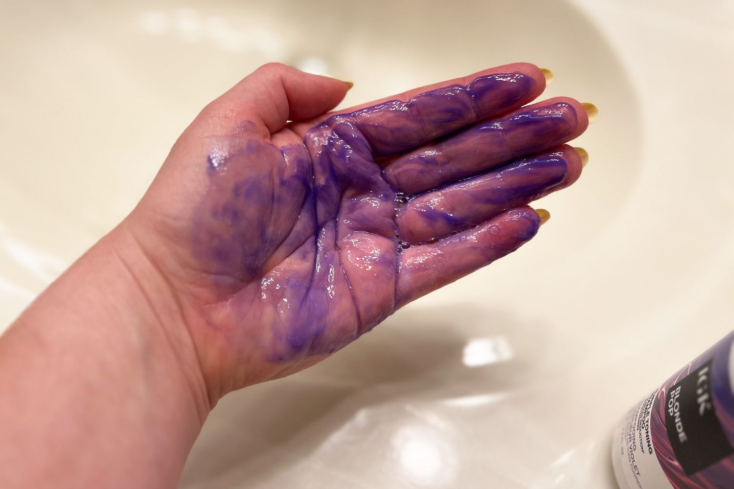 Hand covered with purple IGK Blonde POP Purple Toning Shampoo over a sink 
