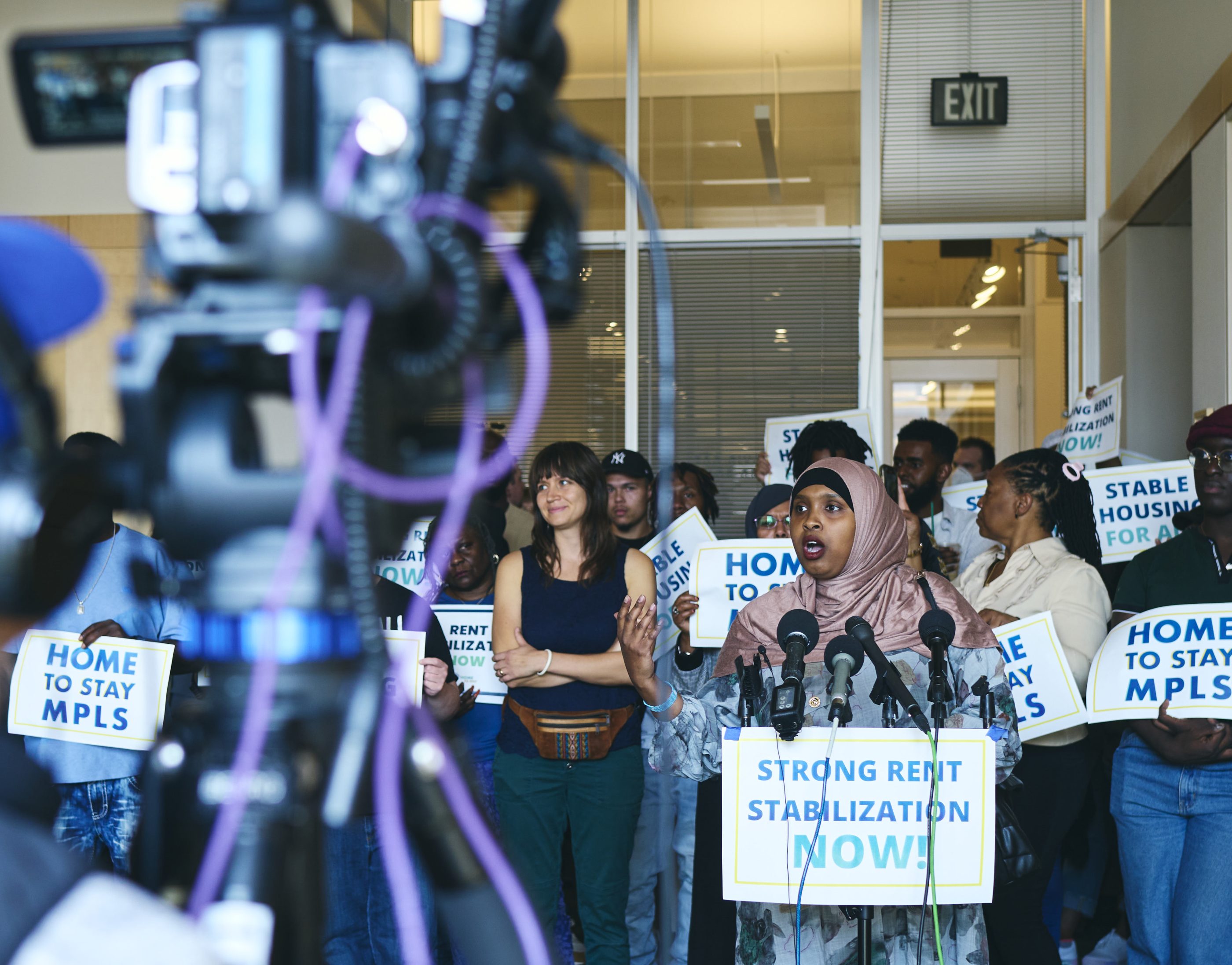 A Black woman in blue flowered dress and dusty pink hijab speaks into several microphones. In foreground, blurry, are news cameras. The woman is part of a large group at a rally, carrying signs promoting rent stabilization and saying "Home to Stay MPLS"