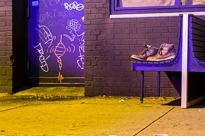 Building exterior and sidewalk lit by neon lights and shoes on a bench and graffiti art on a door