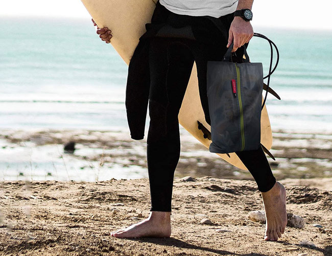 a person wearing black pants ad a white t shirt  holding a surfboard on the beach while carrying a sneaker travel bag in their hand