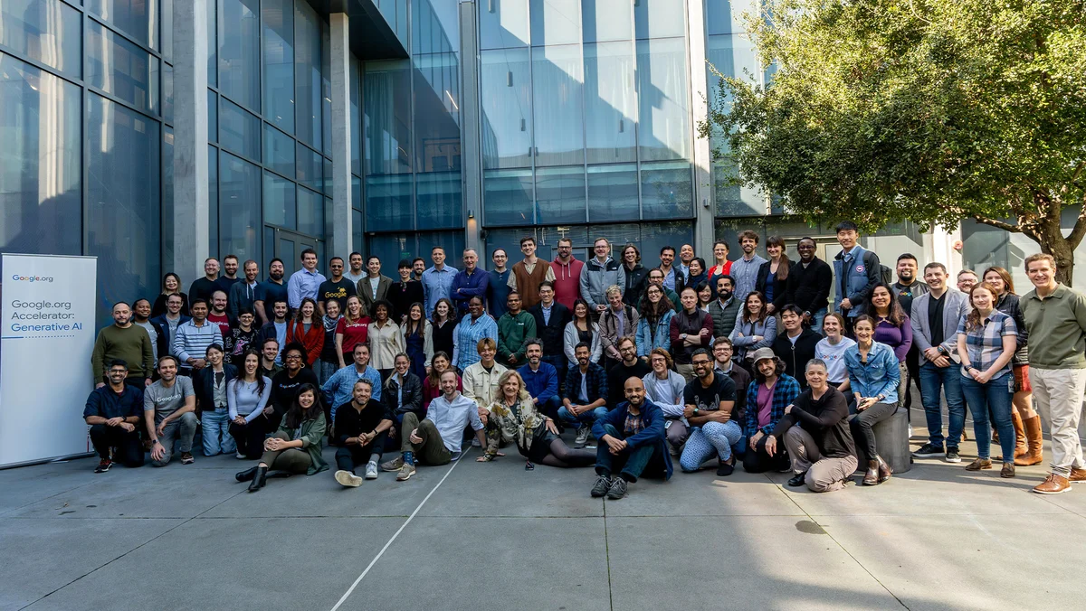 A large group of people stand in front of an office building and a tree in a tight group. A sign reading: Google.org Accelerator: Generative AI stands to the left of the group