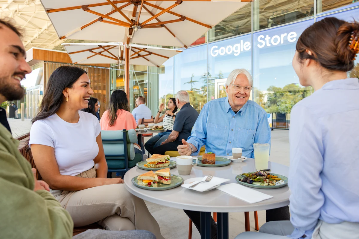 A view of three people eating lunch on a patio with an umbrella over their table, sitting in front of the Google Store entrance.