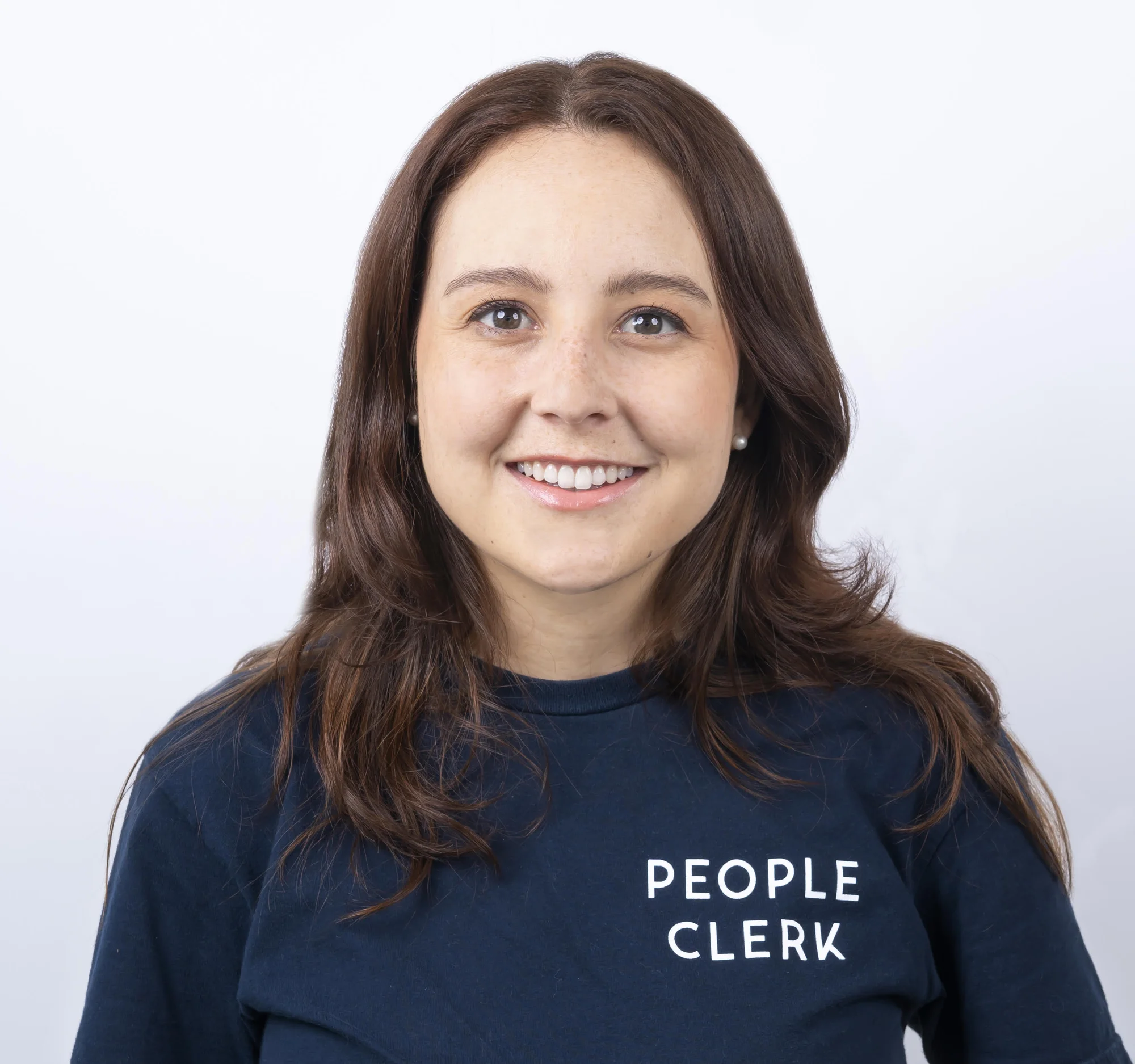A woman with brown hair over her shoulders smiles at camera, wearing a black shirt that says "People Clerk" in white letters.