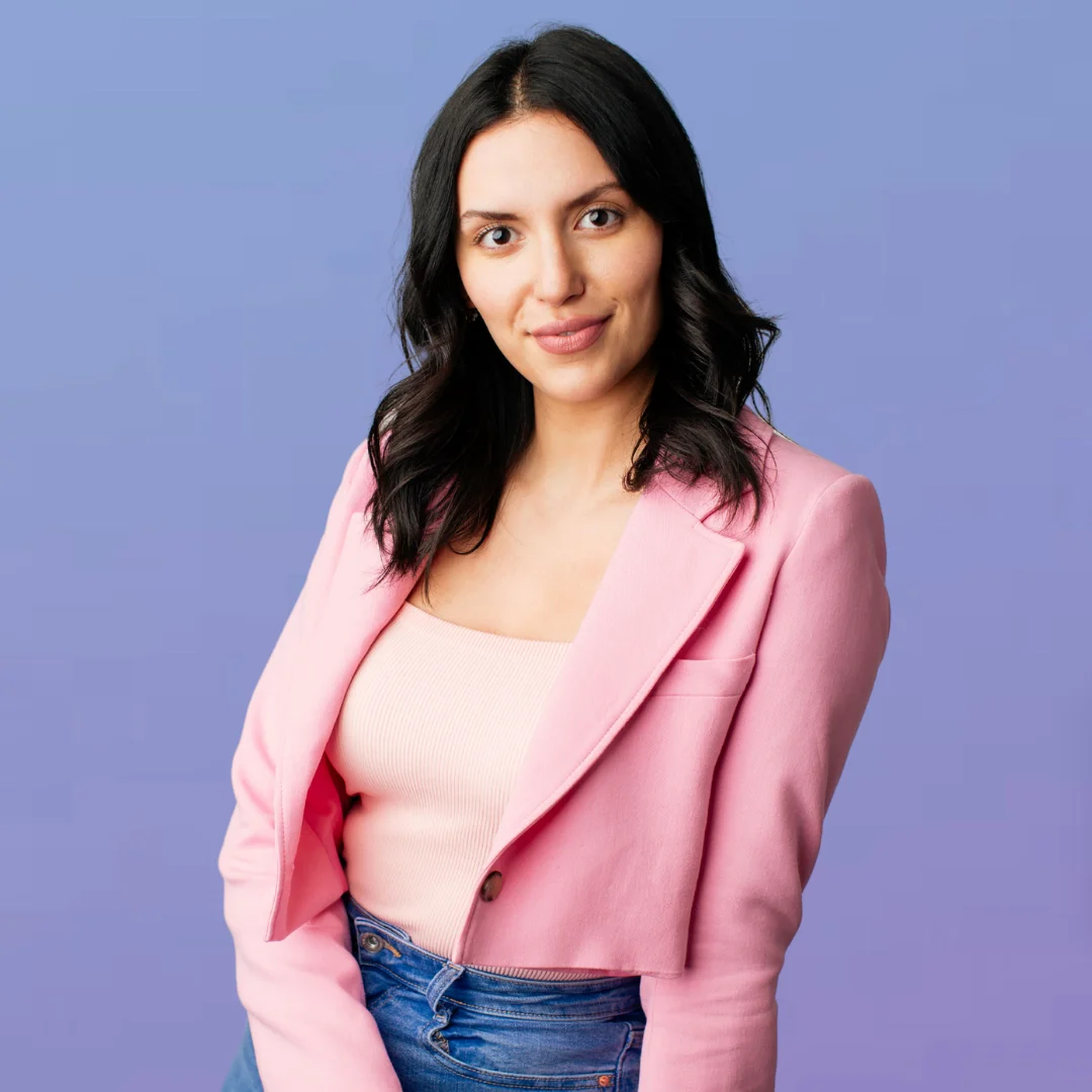 A woman with black hair sits angled towards the camera, wearing a pink blazer and top and blue jeans.