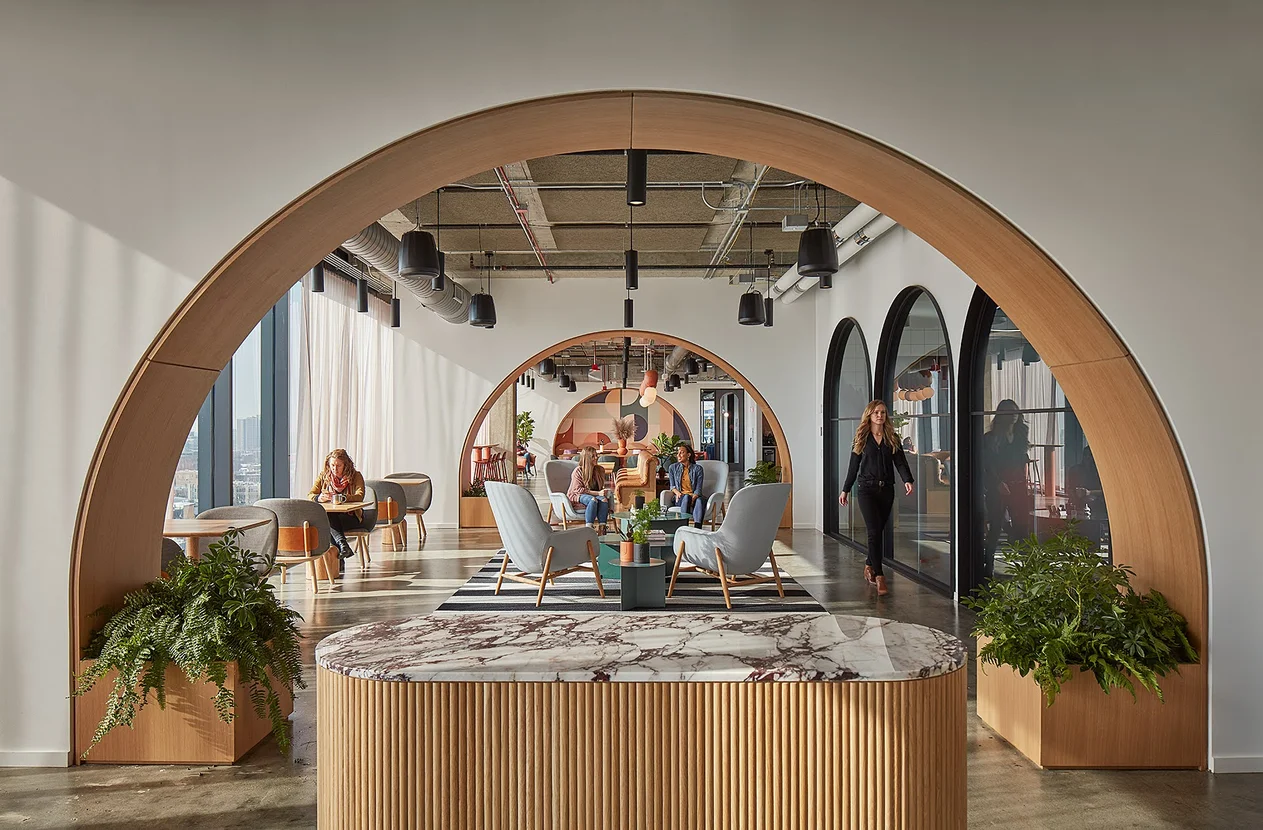 A large archway with four gray chairs in the center, where two Googlers are sitting. Another Googler is sitting at a chair next to large windows overlooking the city. Another employee walks by three arched windows looking out to a hallway.