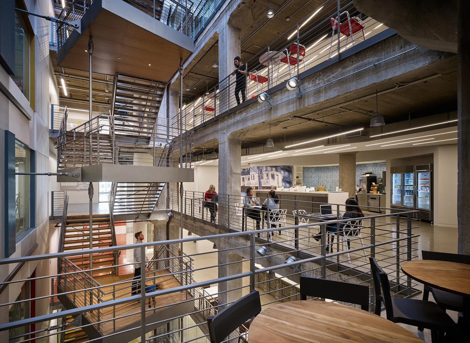 A large staircase leads to three separate floors. A person on the bottom floor looks up towards a kitchen on the next floor, where three Googlers are sitting at tables. A person on the top floor looks down from the balcony.