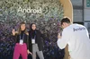 Two MWC attendees pose for a photo in front of a floral backdrop, with a third person taking a photo of them using a Pixel 8 phone.