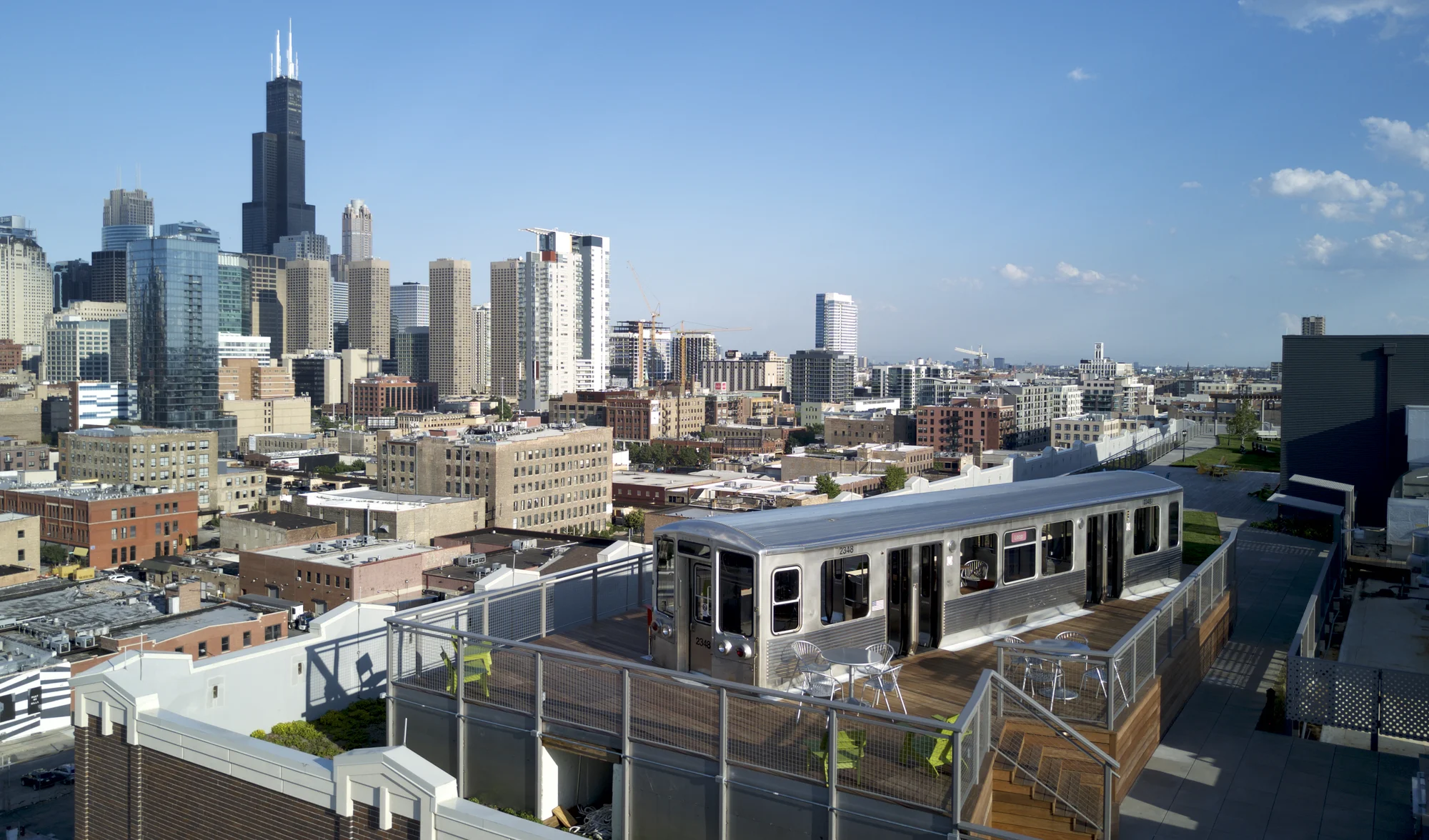 A rooftop overlooking the Chicago skyline with a silver train car. Next to the train are silver and green chairs and two silver tables. Around the rooftop is a balcony wall and a set of stairs that lead down to a second platform.