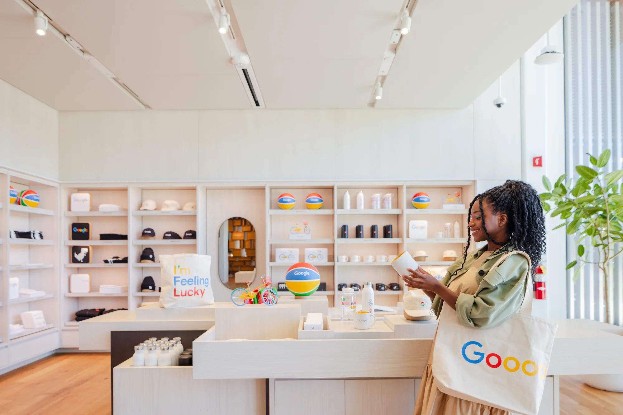 A photo of the interior of the Google Store with a woman standing in front of a display wall and table of Google-branded merchandise. She is holding up a Google-branded mug.