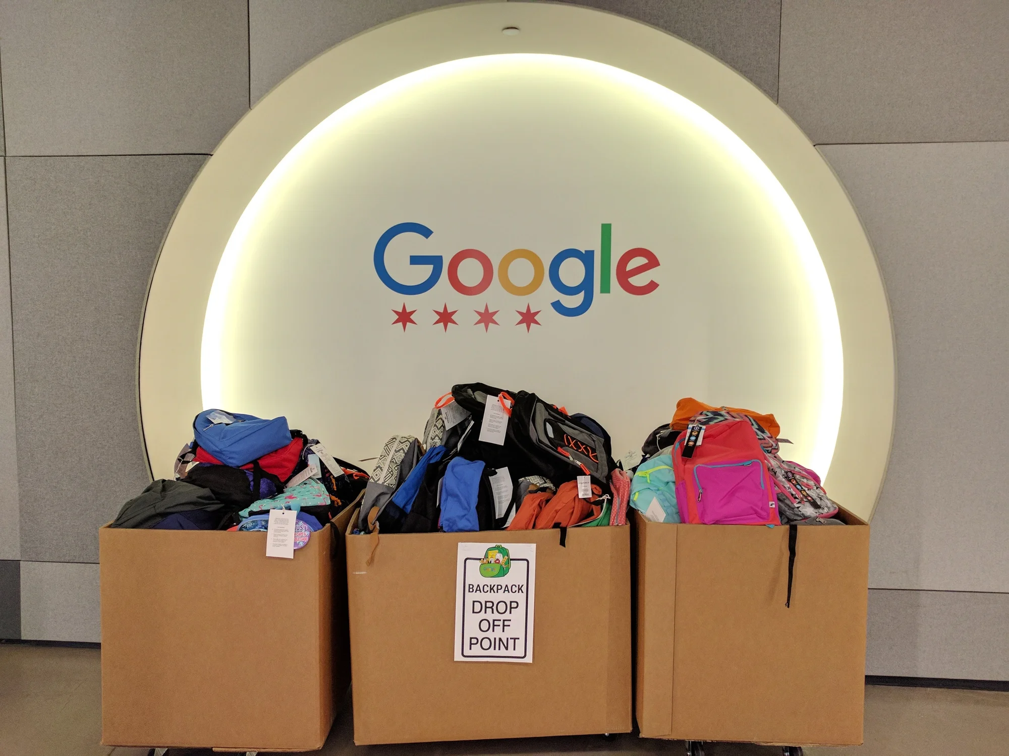 Three large cardboard boxes lined up next to each other, each filled with backpacks of different sizes and colors. On the white wall behind them is the Google logo with four red stars underneath symbolizing the Chicago flag.