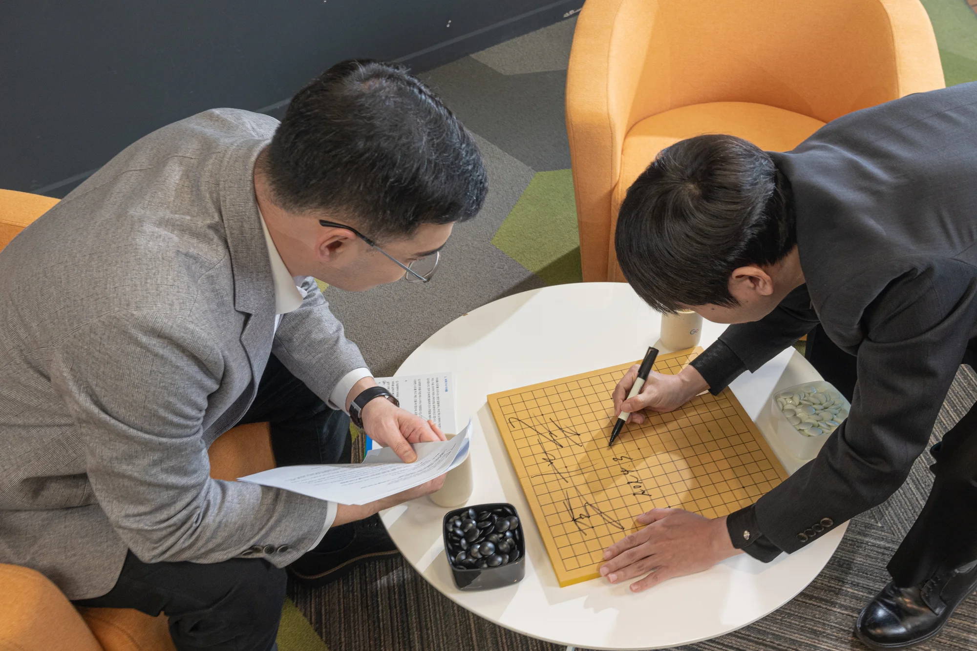 a birds eye view of two people sitting at a white table wearing grey suits. One person holds papers in his hand while the other writes on a wooden square board with a marker.