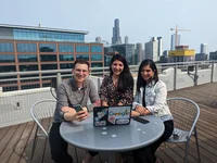 Three Googlers smiling outside at a table. One of the Googlers is holding a phone and the other has a laptop that’s open. The Chicago skyline is in the background.