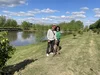 Paula stands with her husband and dog on grass next to a pond on a clear day.