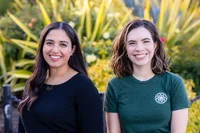 Two women, one wearing a black shirt and one wearing a green shirt, sit side by side facing the camera in front of large green plants.