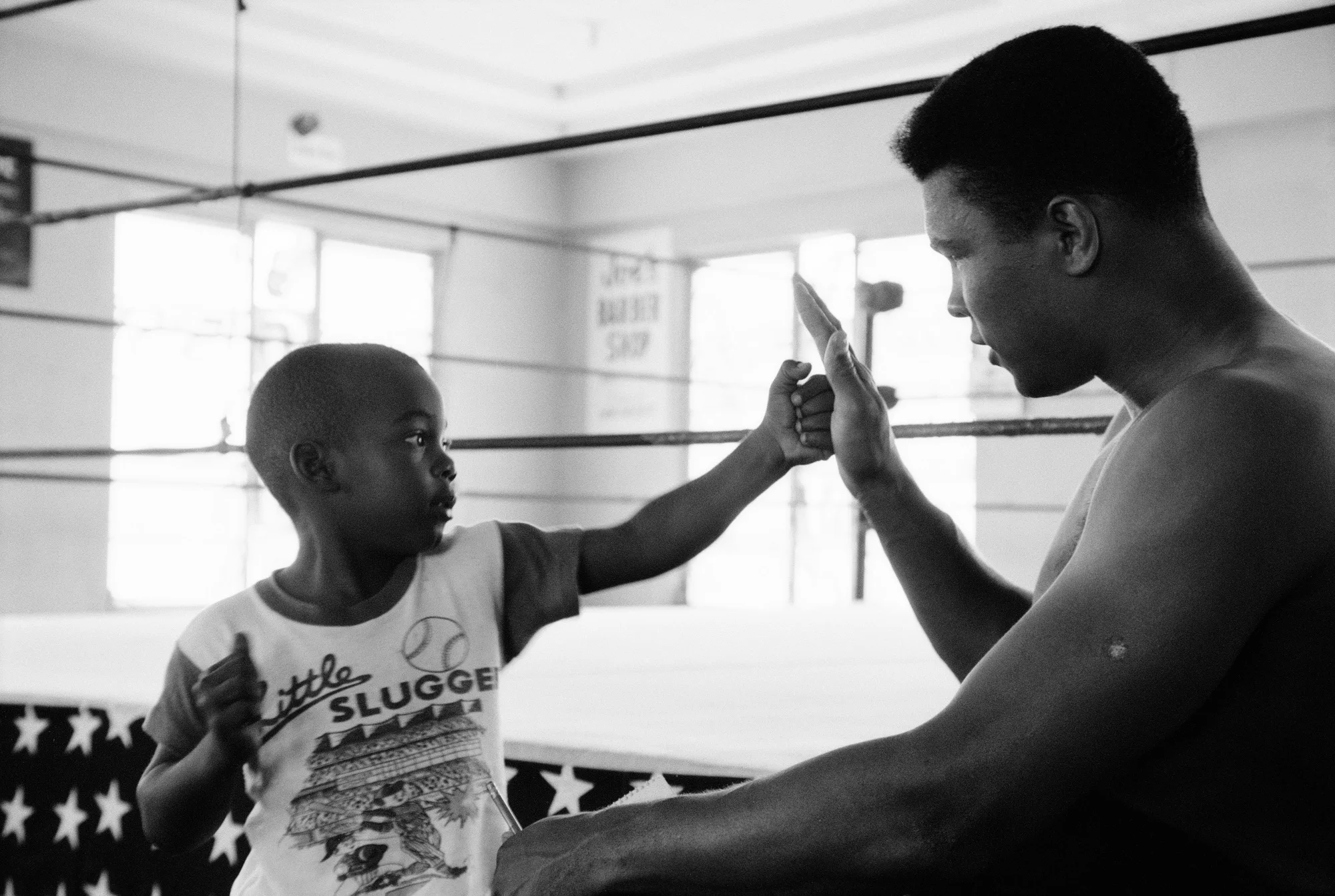 Black-and-white photograph of Muhammed Ali and a small boy, with Ali holding up his hand to block a practice punch from the child.