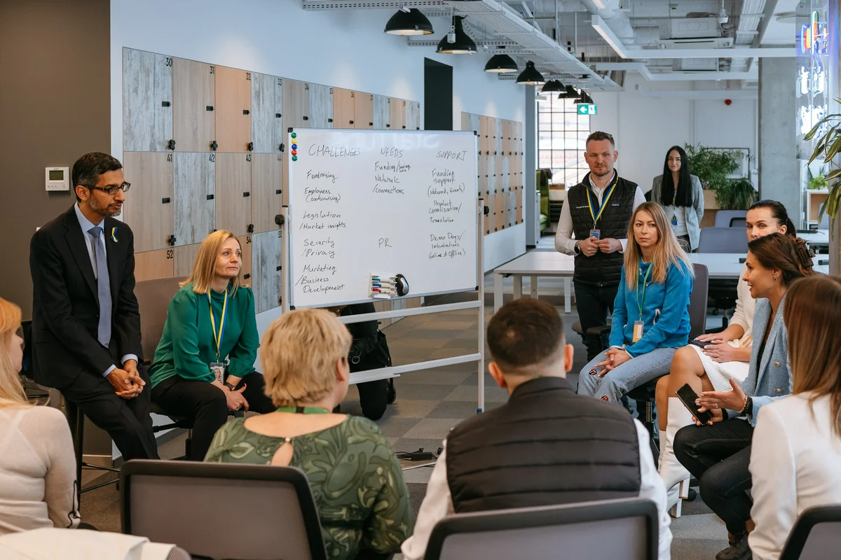 Google CEO Sundar Pichai sits on a stool to the left of a group of NGO employees in a circle at a Google’s startup campus in Warsaw. He is listening to a woman at the far right of the group. There is a whiteboard in the center of the photo.