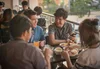A group of four young adults are having a meal at an alfresco restaurant. One of them is a bespectacled boy who is holding a smartphone in his hands and chatting with his friend.