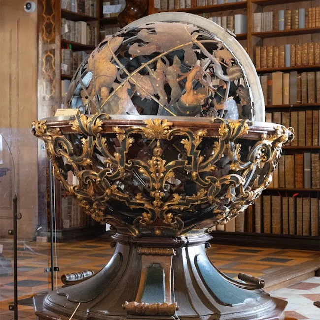 An ornate celestial globe with intricate design, displayed in a library with bookshelves in the background.