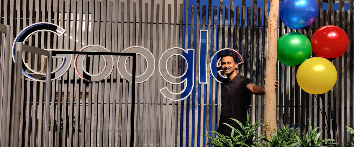 Johnson standing in front of a large, wooden Google sign. He leans on a tree. There is a set of blue, green, red, and yellow balloons next to him.