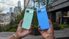 Two hands holding Pixel 8a devices; one is bright blue and the other is a mint green. The backs of the phone face the camera. A city scape is in the background.