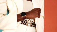 A woman in a white pantsuit and geometric belt modeling the new Brother Vellies woven leather accessory band in oak for Fitbit Sense 2 and Versa 4.