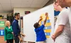 Sundar and 4-H President and CEO Jennifer Sirangelo stand to the left of a whiteboard watching a student in a blue top reaching up to complete a coding activity. Two other students in white look on from the right of the whiteboard.