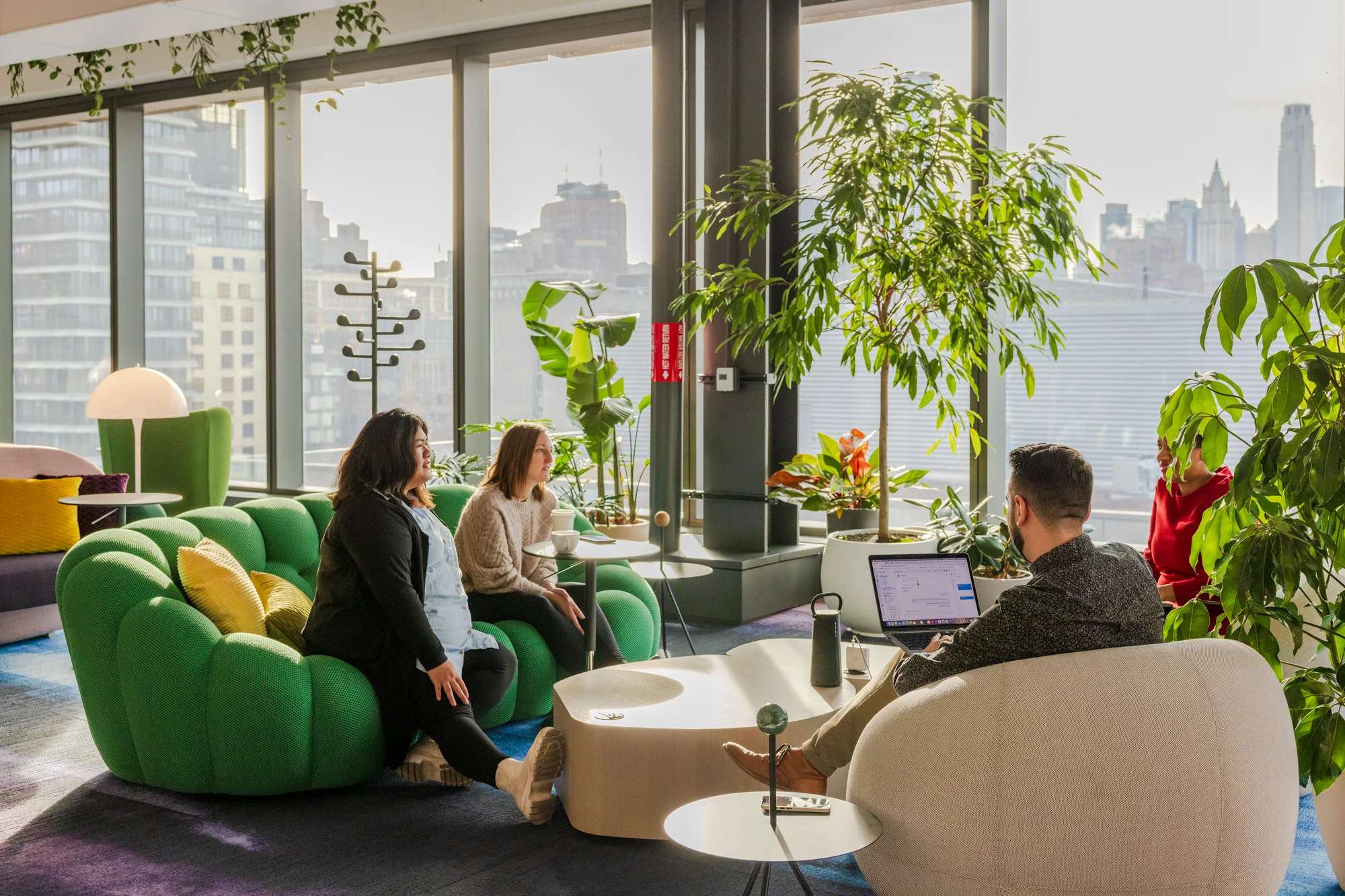 Two Googlers sit on a green couch, collaborating with their colleagues in one of the building's work lounges. In the background, floor to ceiling windows look out upon the New York skyline.