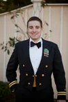 Kevin Ryan stands, looking into the camera and smiling, wearing his military dress jacket with a white button-up shit and black bow tie.