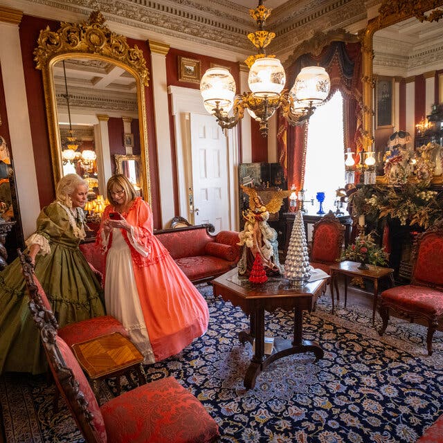 Two women dressed in long, flowing Civil War-era dresses stand in an Antebellum home containing ornate furniture and large decorative mirrors. 