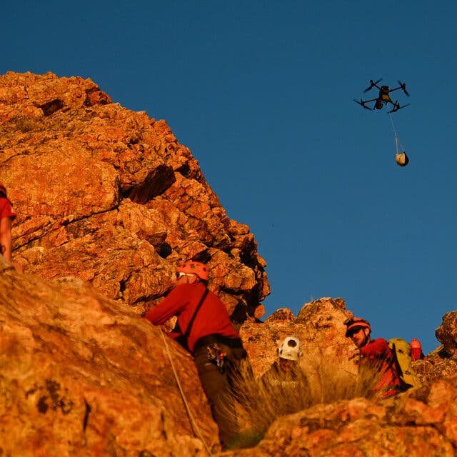 A drone carrying supplies flies above people wearing red jackets and orange helmets who are standing in a rocky landscape.